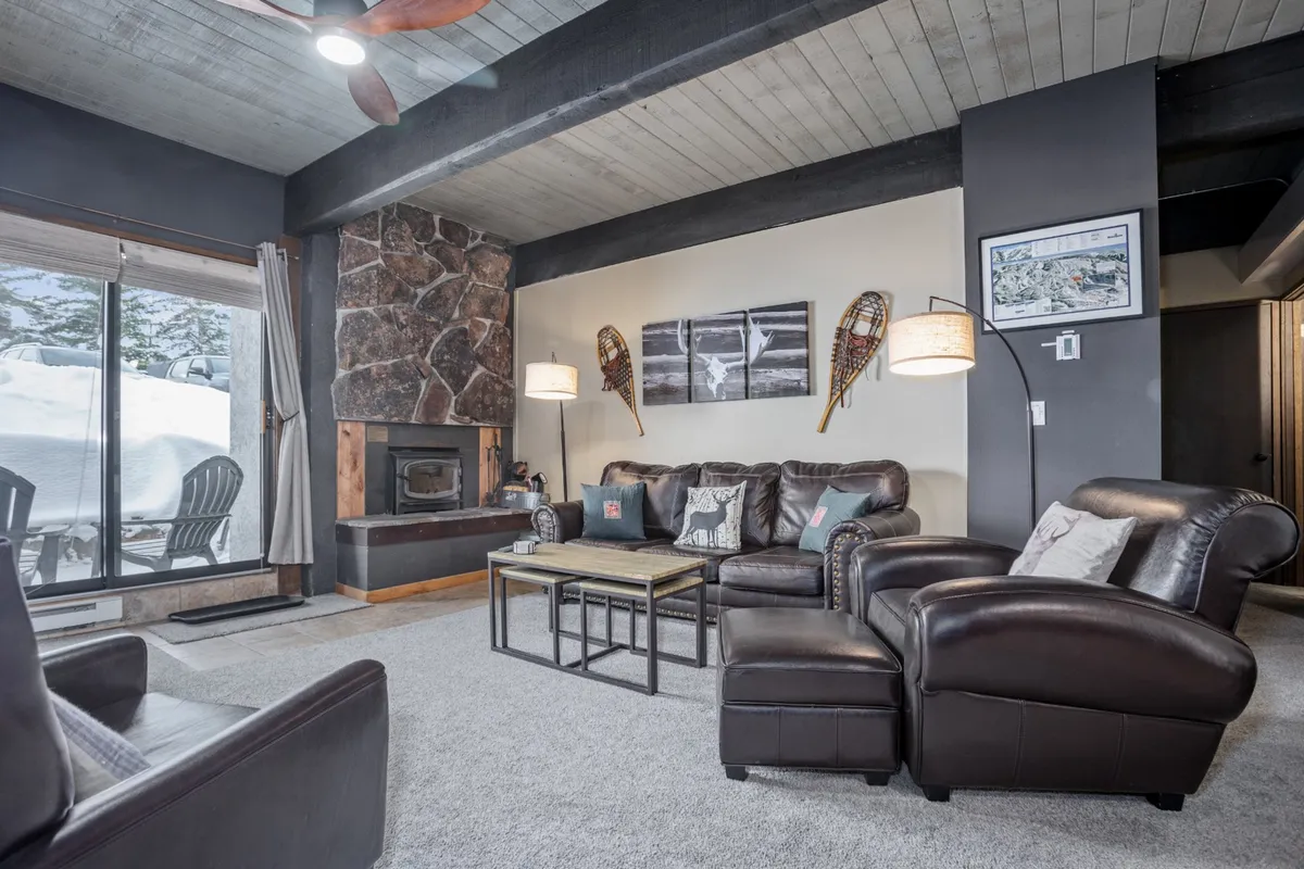 A spacious living room with a striking stone fireplace, plush leather sofas, large windows letting in natural light, and tasteful artwork on the walls. The setting is ideal for relaxing, socializing, or enjoying a warm beverage by the fire.