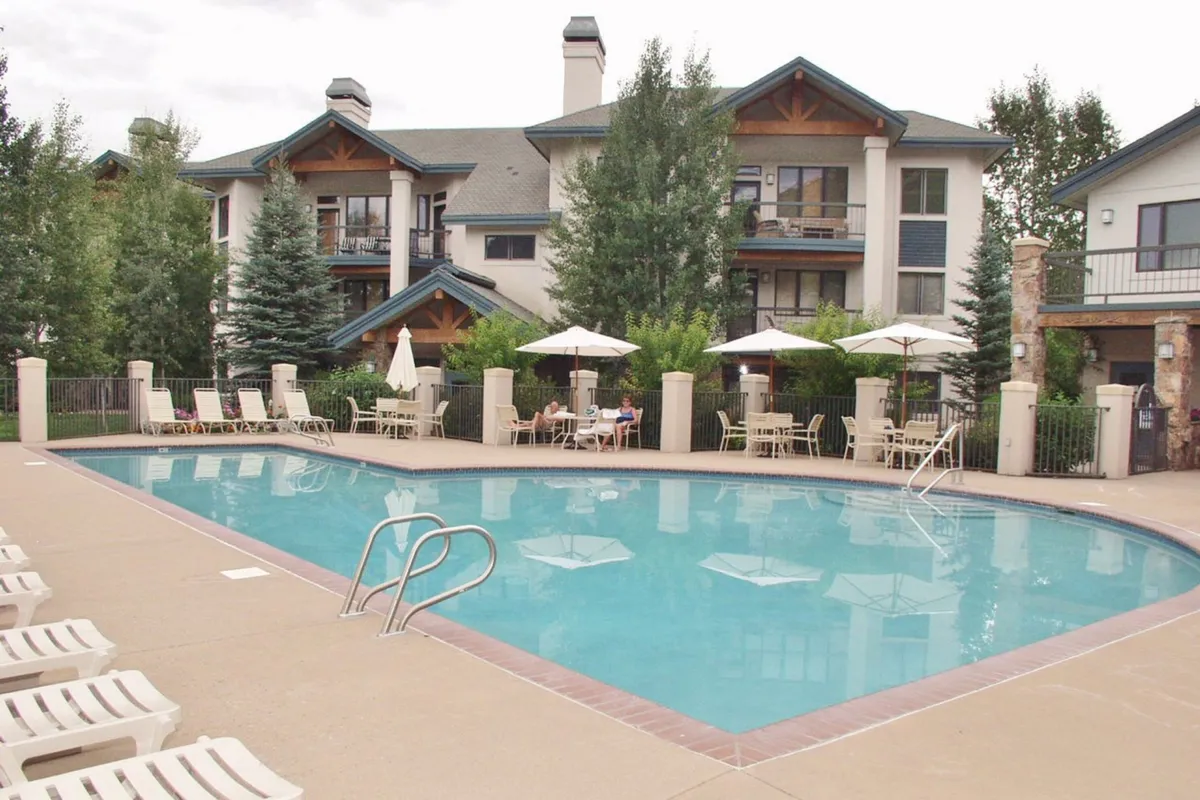 This image features a lively exterior setting with a swimming pool surrounded by lounge chairs and patio umbrellas, set against the facade of the building. The scene suggests relaxation and social activities like swimming, sunbathing, and enjoying refreshments after a day outdoors.