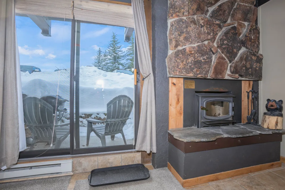 A living area with a stone fireplace, large glass sliding door, and outdoor chairs visible through the window. It bridges the indoor comfort with scenic outdoor views, highlighting the private patio's easy access.