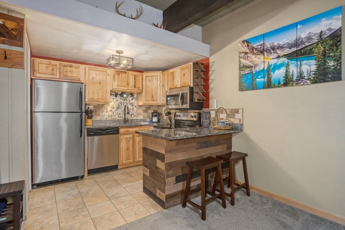 A well-appointed kitchen with a granite countertop, wooden cabinetry, a mountain landscape painting, and bar stools. The space is warm and designed for both cooking and socializing, fitting seamlessly into the open-concept layout.