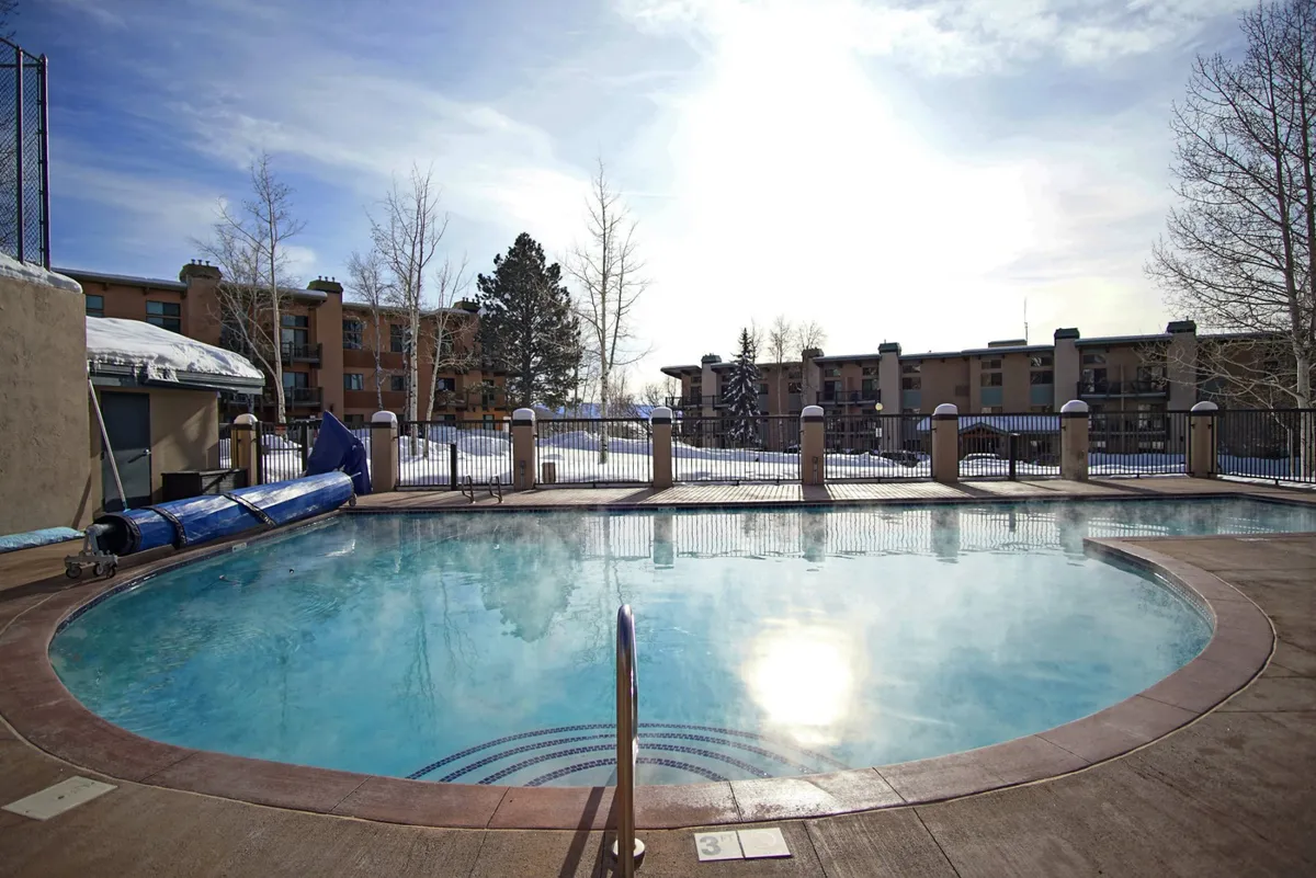 A tranquil, heated swimming pool is surrounded by snow with residential buildings in the background. Steam rises from the water, suggesting warmth and relaxation despite the wintry setting.