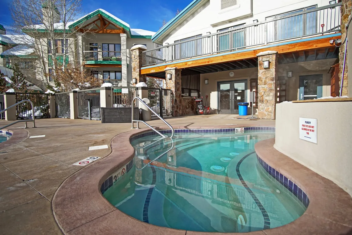 A hot tub is set on a wooden deck bordered by a stone wall, with steam rising from the water. Snow-covered trees and a tranquil winter landscape frame the space, suggesting an inviting spot to unwind after outdoor adventures.