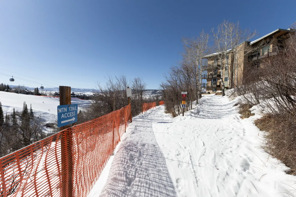 A snow-covered path leads from the building to a ski trail, with a cable car overhead and signage indicating trail access. The surrounding area is blanketed in fresh snow, and a building is visible in the background, creating a clear connection between the condo and ski terrain.