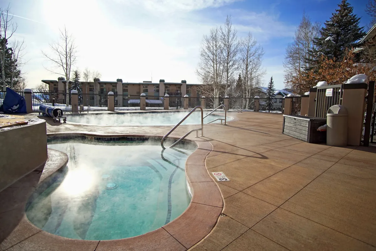 An inviting outdoor hot tub surrounded by a fenced deck, with steam rising from the water and mature trees in the background. The area feels private and tranquil, offering a relaxing spot no matter the season.