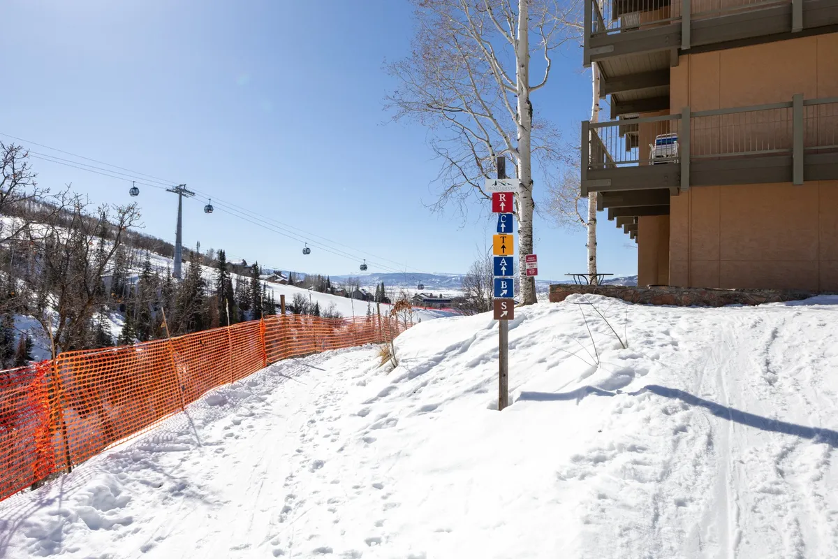 The image displays the exterior of the property, nestled in a snowy landscape with a clear view of the building, signage, and a ski lift in the background. The scene includes snow-covered ground and people enjoying the outdoors, highlighting direct access to mountain activities and a sense of place.