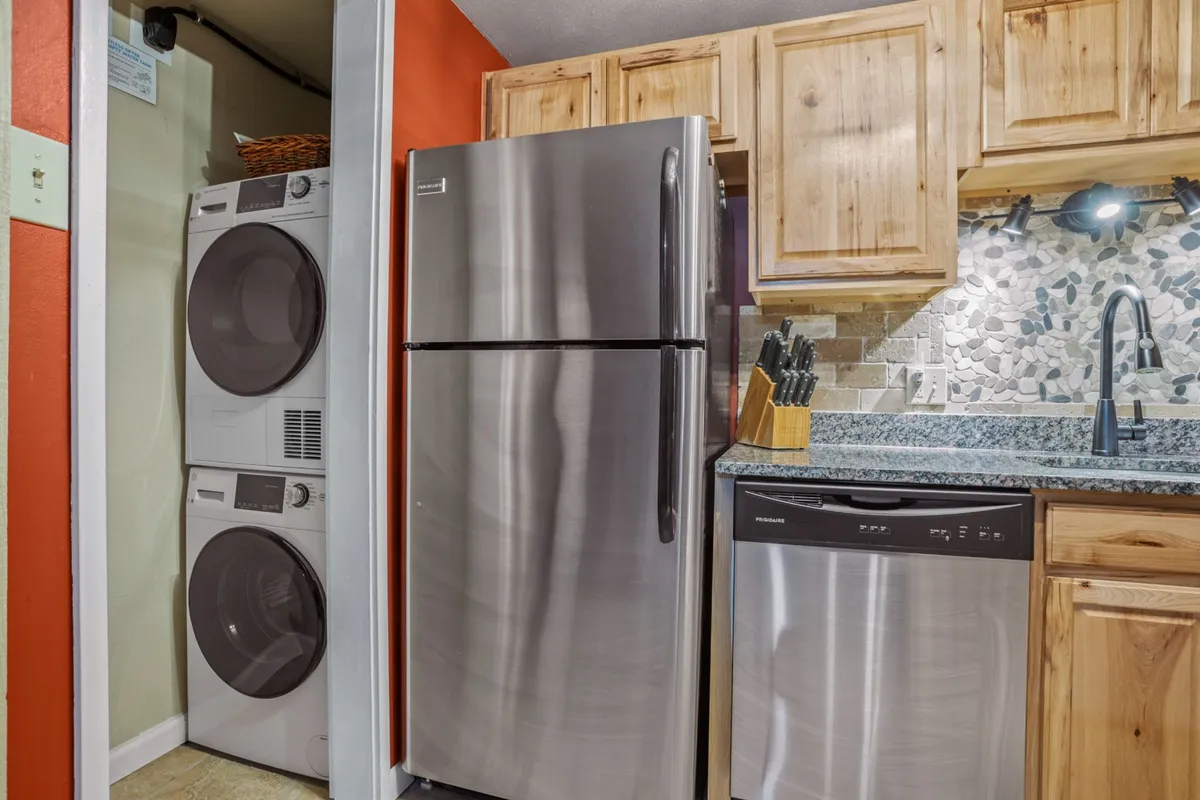 This image captures a multi-functional interior space featuring stainless steel appliances, a granite countertop, colorful wall paint, and wooden cabinets. The composition subtly includes both kitchen and laundry appliances, suggesting the convenience of multitasking laundry and meal prep in one inviting area.